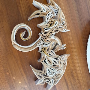 May include: A dragon-shaped sculpture made from folded pages of a book. The sculpture is made of cream and brown paper and is displayed on a wooden surface.