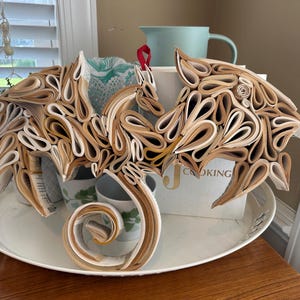 May include: A dragon sculpture made from rolled pages of a book. The dragon is brown and white and has its wings spread out. It is sitting on a white tray with a white background.