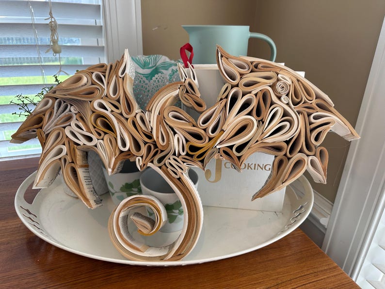 May include: A dragon sculpture made from rolled pages of a book. The dragon is brown and white and has its wings spread out. It is sitting on a white tray with a white background.