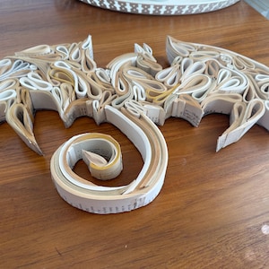 May include: A dragon sculpture made from rolled up pages of a book. The dragon is white and cream colored and has a curled tail.