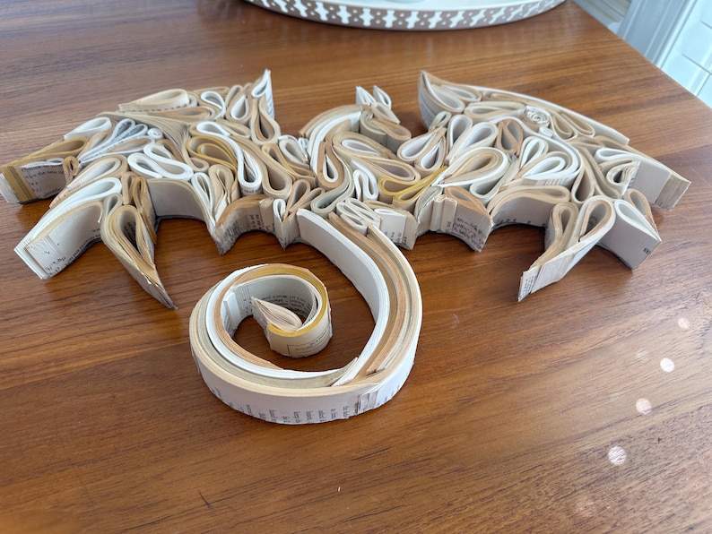 May include: A dragon sculpture made from rolled up pages of a book. The dragon is white and cream colored and has a curled tail.