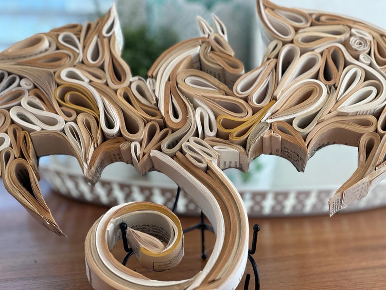 May include: A dragon sculpture made from rolled pages of a book. The dragon is brown and white with a curled tail. The sculpture is on a black metal stand.