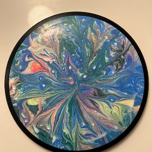 May include: Round abstract art piece with a black border. The artwork features a swirling pattern of blues, greens, pinks, and yellows, creating a dynamic and colorful design. The piece is a decorative item, suitable for wall display.