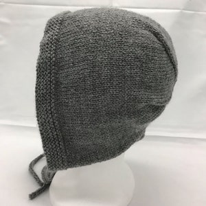 May include: Gray knitted baby bonnet with a tie. The bonnet is made of a soft, textured yarn and has a simple, classic design.