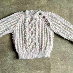 May include: A white, hand-knitted sweater with a textured stitch pattern and cable knit detailing. The sweater has long sleeves and is adorned with three brown buttons on the shoulder. It features subtle flecks of pastel colors throughout.