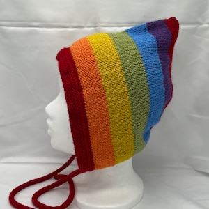 May include: A rainbow striped knitted hat with a pointed top and red ties. The hat is displayed on a white mannequin head.