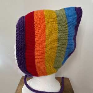 May include: A rainbow striped knitted hat with a purple tie. The hat is made of soft yarn and has a warm, cozy feel.