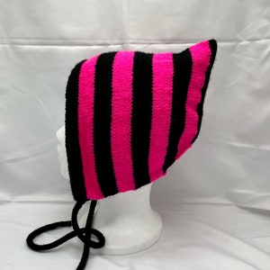 May include: A pink and black striped knit hat with a long black tie. The hat has a pointed top and is designed to cover the ears.