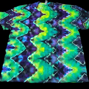 Op de afbeelding: Een tie-dye t-shirt met een levendig zigzagpatroon. Het shirt heeft een mix van groene, blauwe, paarse en gele tinten. Het shirt met korte mouwen wordt gepresenteerd tegen een zwarte achtergrond.