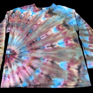 Op de afbeelding: Een lange mouwen tie-dye shirt met een spiraalpatroon. Het shirt heeft een mix van kleuren, waaronder blauw, roze, bruin en grijs. De mouwen hebben geribde manchetten en het shirt ligt op een zwarte achtergrond.