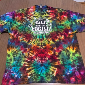 Puede incluir: Una camiseta tie-dye con un patrón de espiral colorido. La camiseta tiene un gráfico blanco con el texto "I'm Bill and it's a Thrill!" con un banjo, una guitarra y otros instrumentos musicales.