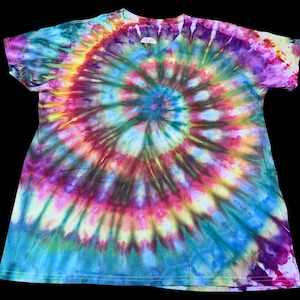Op de afbeelding: Een tie-dye V-hals T-shirt met een spiraalpatroon in tinten blauw, paars, geel en groen. Het shirt heeft korte mouwen en een relaxte pasvorm, perfect voor casual wear. Het kleurrijke ontwerp is opvallend.