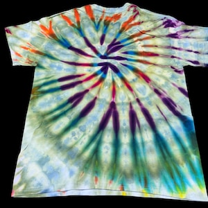 Op de afbeelding: Een tie-dye t-shirt met een spiraalvormig ontwerp. Het shirt heeft een mix van kleuren, waaronder oranje, rood, paars, blauw en groen, die vanuit het midden uitstralen. Het shirt is gemaakt van een zachte, lichtgekleurde stof.