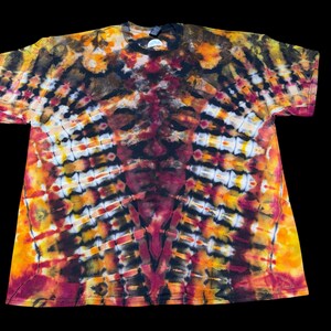 Op de afbeelding: Een tie-dye t-shirt in een levendige mix van kleuren, waaronder oranje, geel, bruin en bordeauxrood. Het ontwerp heeft een symmetrisch patroon met een centraal gezichtachtig beeld. Het shirt met korte mouwen staat tegen een zwarte achtergrond.