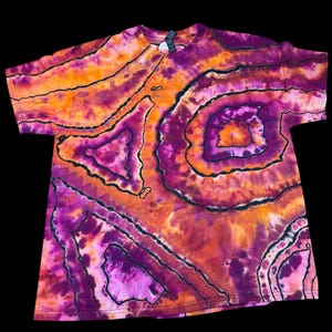 Op de afbeelding: Een tie-dye t-shirt in tinten oranje, paars en roze, met zwarte omtrekdetails. Het shirt heeft een ronde hals en korte mouwen. Het tie-dye patroon creëert een gelaagd, cirkelvormig ontwerp.