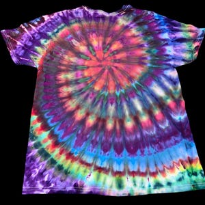 Op de afbeelding: Een t-shirt met korte mouwen en een levendig tie-dye patroon. Het ontwerp heeft een spiraal van kleuren, waaronder paars, roze, blauw, groen en geel. Het shirt ligt plat op een zwarte achtergrond, wat het ingewikkelde verfwerk laat zien.
