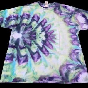 Op de afbeelding: Een tie-dye t-shirt in tinten paars, blauw en groen. Het shirt heeft een symmetrisch bloemenpatroon in het midden, waarbij de kleuren over de stof mengen en wervelen. Het t-shirt is een ronde hals stijl.
