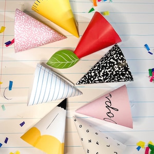 Könnte beinhalten: Eine Sammlung bunter Partyhüte aus Papier in verschiedenen Mustern und Designs, darunter ein gelbes Lineal, rosa Blumenmuster, rot, ein schwarz-weißes Notizbuch, blaue Streifen, ein Bleistift, ein rosa Hut mit dem Wort "pops" und ein weißer Hut mit Buchstaben. Konfetti ist um die Hüte verstreut.