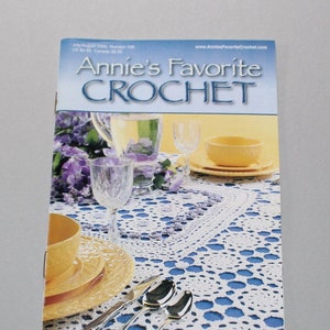 May include: A magazine cover for Annie's Favorite Crochet, featuring a white crocheted tablecloth with a blue background, set with yellow dishes, silverware, and a pitcher of water. The magazine cover text reads "Annie's Favorite Crochet" and "July/August 2000, Number 100".