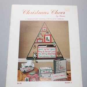 May include: A vintage book cover titled "Christmas Cheer" by Ninna, featuring needlepoint designs. The cover displays a Christmas tree-shaped display with various needlepoint projects, including boxes and ornaments. The text "Year after Year May it never fail the JOY of Sharing Christmas Mail" is visible.