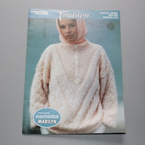 May include: A pink knitted sweater with a zipper and a cable knit pattern. The sweater is modeled by a woman wearing a pink headscarf. The text "Leisure Arts Fashion Leaflet 1308 $2.50 MARILYN featuring nomotta MARILYN" is printed on the page.