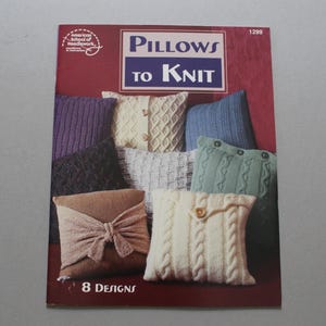 May include: A softcover instructional booklet titled Pillows to Knit, number 1299, featuring 8 designs. The cover displays a collection of various knitted pillows in colors including cream, blue, green, brown, and black, with button and bow details.