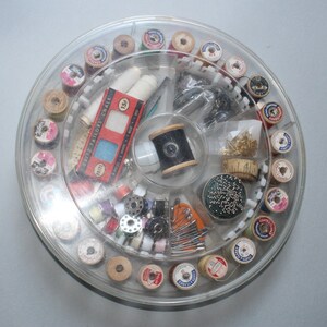 May include: A clear plastic sewing kit with a rotating circular compartment containing spools of thread, buttons, needles, and other sewing supplies. The kit also includes a small box of tailor's chalk.