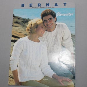 May include: A knitting pattern book cover featuring a couple wearing white cable knit sweaters. The book is titled "Bernat Gloucester" and has the book number 573. The price is $2.75 in the US and $3.30 in Canada.