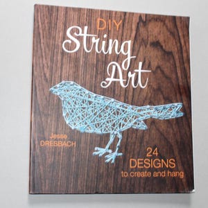 May include: A book cover with a wood grain effect background displays the title "DIY String Art" in white script. A blue string art bird is shown. The book is by Jesse Dresbach and includes 24 designs to create and hang.