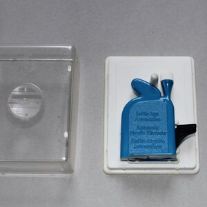 May include: A blue Infila Age Automatic Needle Threader, with text, sits in a white tray, next to a clear plastic case. The threader has a black lever and white accents.