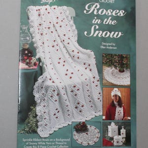 May include: A book cover featuring a white crochet blanket with red rose accents, titled "Roses in the Snow." The cover also shows a matching hat, scarf, and other crochet items. The text reads "The Needlecraft Shop."