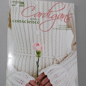 May include: Book cover with a white knitted cardigan and a pink flower. The title is "Cardigans with a conscience" and the subtitle is "6 Knit Designs in Sizes Small to 3X by Melissa Leapman".