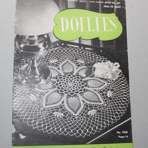 May include: A vintage book cover for a collection of doilies with a black and white photo of a crocheted doily on a table. The book cover is green and white with the title "Doilies" and the text "A Collection of Fascinating New Designs".