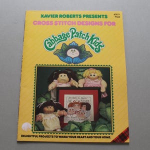 May include: A yellow softcover book titled Xavier Roberts Presents Cross Stitch Designs for Cabbage Patch Kids #7677 Plaid. The cover features three dolls and a framed cross stitch sampler with text, surrounded by green cabbage leaf illustrations.