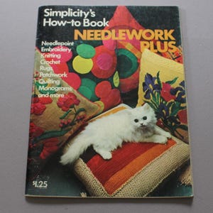 May include: A softcover instructional book titled Simplicity's How-to Book NEEDLEWORK PLUS. The cover features a white cat resting on a striped cushion, surrounded by various needlework projects including floral pillows and colorful circular patterns.