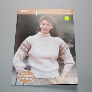 May include: A white knitted sweater with a turtleneck and brown, blue, and white striped sleeves. The sweater is featured in a fashion magazine with the text "Leisure Arts Fashion Leaflet 1308 $2.50 SUPER-SHOW featuring nomotta SUPER-SHOW".