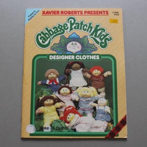 May include: A Cabbage Patch Kids Designer Clothes pattern booklet by Xavier Roberts featuring illustrations of dolls in various outfits. The cover includes the text "Cabbage Patch Kids," "Designer Clothes," and "Make 25 Outfits" with a plaid accent.