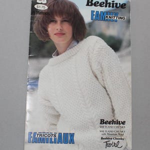 May include: A knitting pattern booklet featuring a woman wearing a white cable knit jumper. The booklet is titled "Beehive Family Knitting" and includes the text "Beehive Shetland Chunky Twirl".