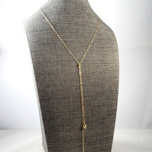 May include: A gold chain necklace with two small gold balls hanging from the bottom. The necklace is displayed on a gray mannequin.