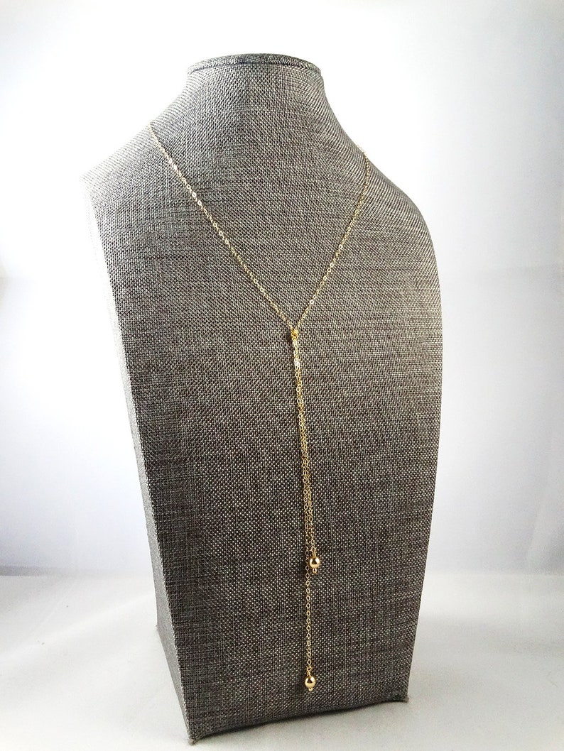 May include: A gold chain necklace with two small gold balls hanging from the bottom. The necklace is displayed on a gray mannequin.
