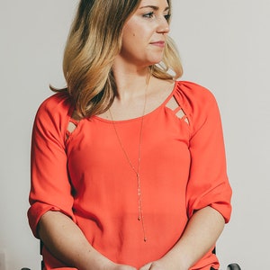 May include: A woman in a bright orange top with a gold necklace sits in a wheelchair. She is looking to the right of the frame.