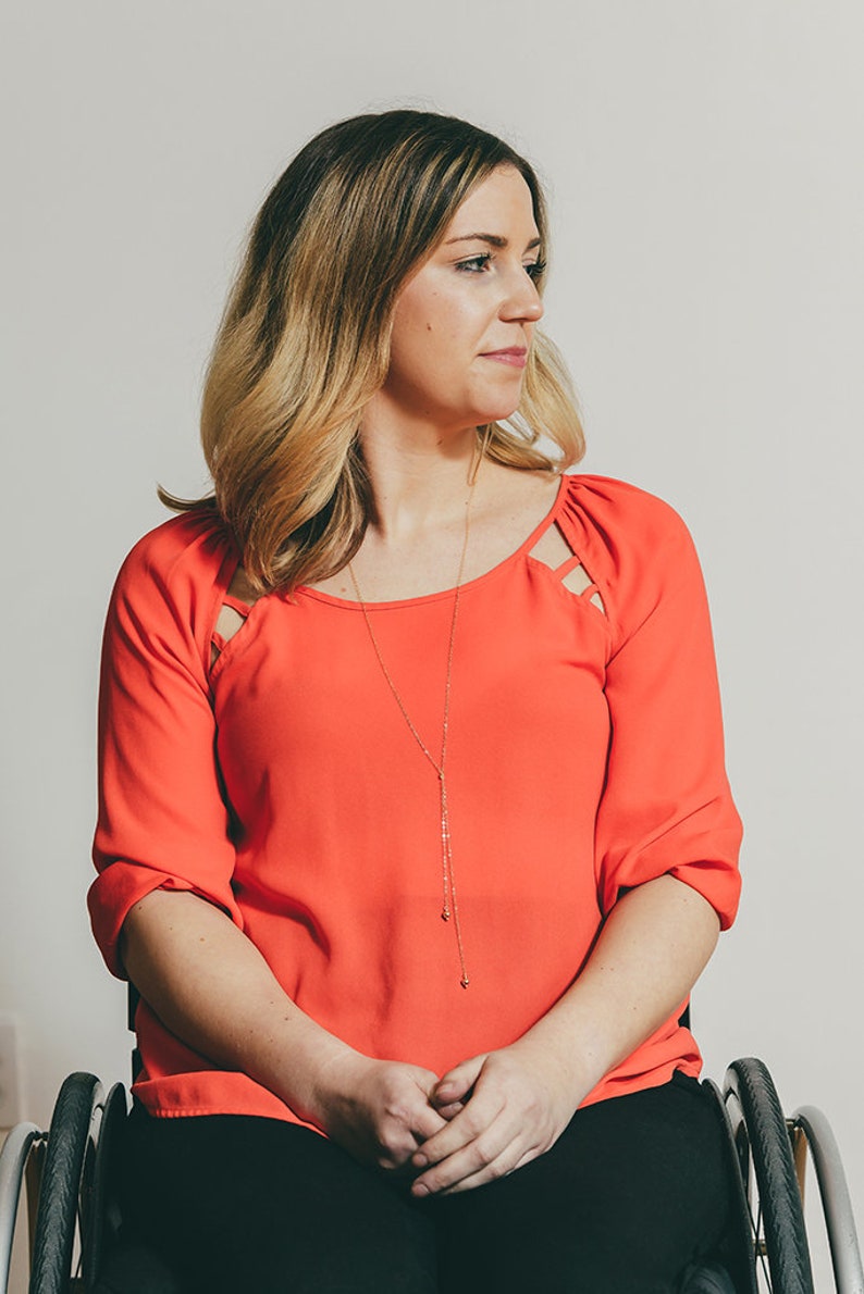 May include: A woman in a bright orange top with a gold necklace sits in a wheelchair. She is looking to the right of the frame.