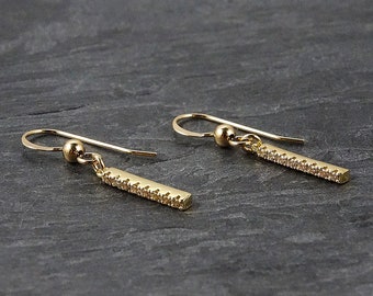 May include: A pair of gold dangle earrings with a small, round bead at the top and a thin, rectangular bar with small, clear stones set into it.