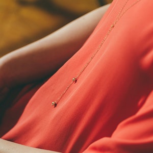 May include: A delicate gold chain necklace with two small round beads hanging from it. The necklace is worn by a person wearing a red shirt.