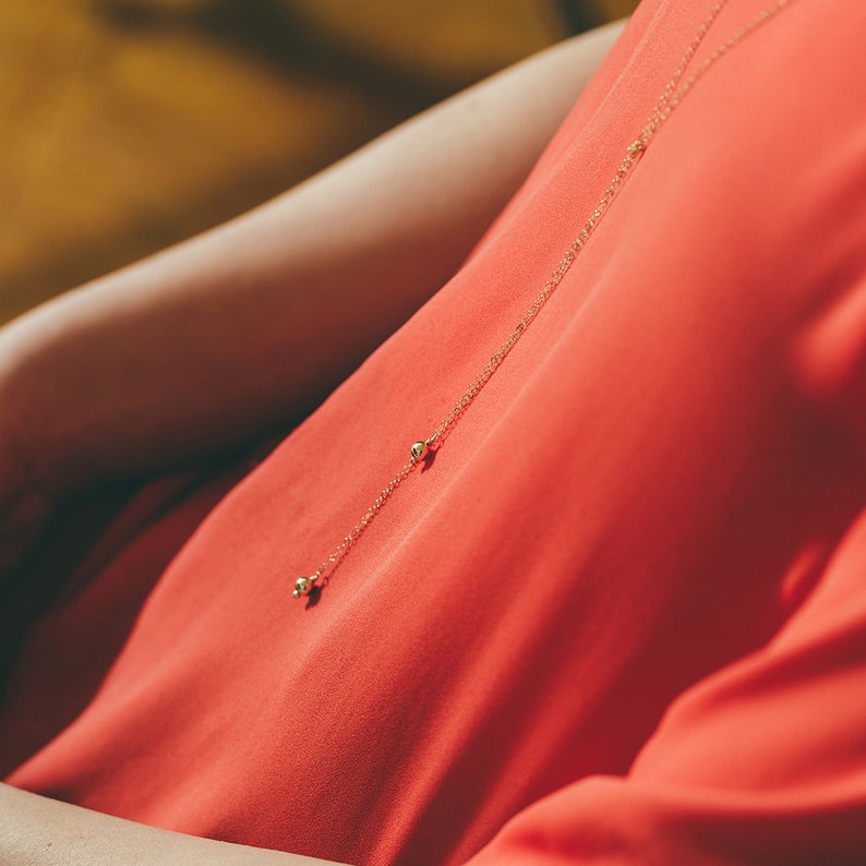 May include: A delicate gold chain necklace with two small round beads hanging from it. The necklace is worn by a person wearing a red shirt.