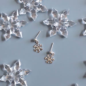May include: A pair of silver stud earrings featuring small, dangling snowflake charms with a polished finish. The earrings are positioned among several larger, clear, translucent plastic snowflake decorations on a solid light grey surface.