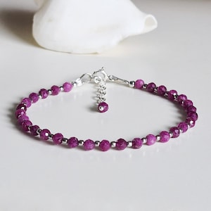 May include: A delicate bracelet featuring small, faceted magenta-colored beads alternating with tiny, smooth silver-toned metal spheres. The piece includes a silver-toned metal lobster claw clasp and a short extender chain finished with a single bead.