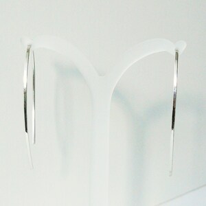 May include: A pair of silver hoop earrings with a thin, flat design. The earrings are hanging from a white display stand.