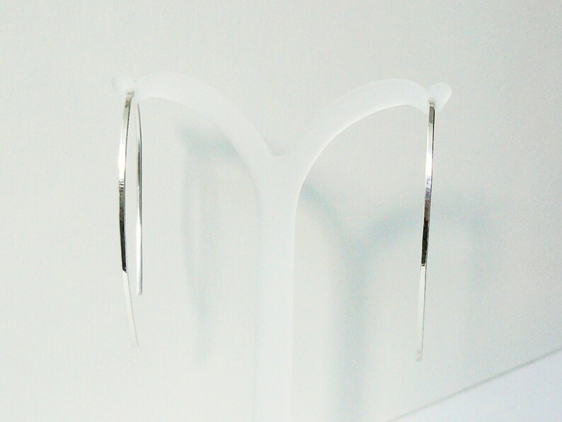 May include: A pair of silver hoop earrings with a thin, flat design. The earrings are hanging from a white display stand.