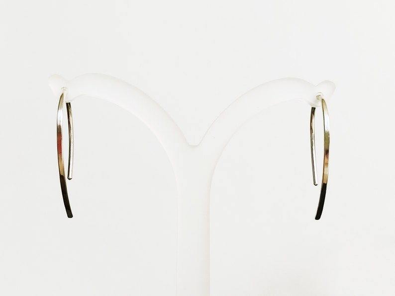 May include: A pair of silver hoop earrings with a simple, curved design. The earrings are displayed on a white stand.